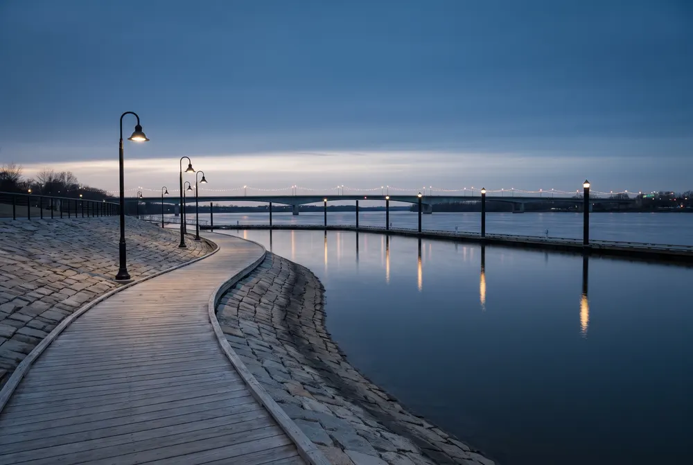 Illustration générée : une promenade de bois le long d'un fleuve à l'heure bleue, des lampadaires noirs et, au loin, un long pont éclairé